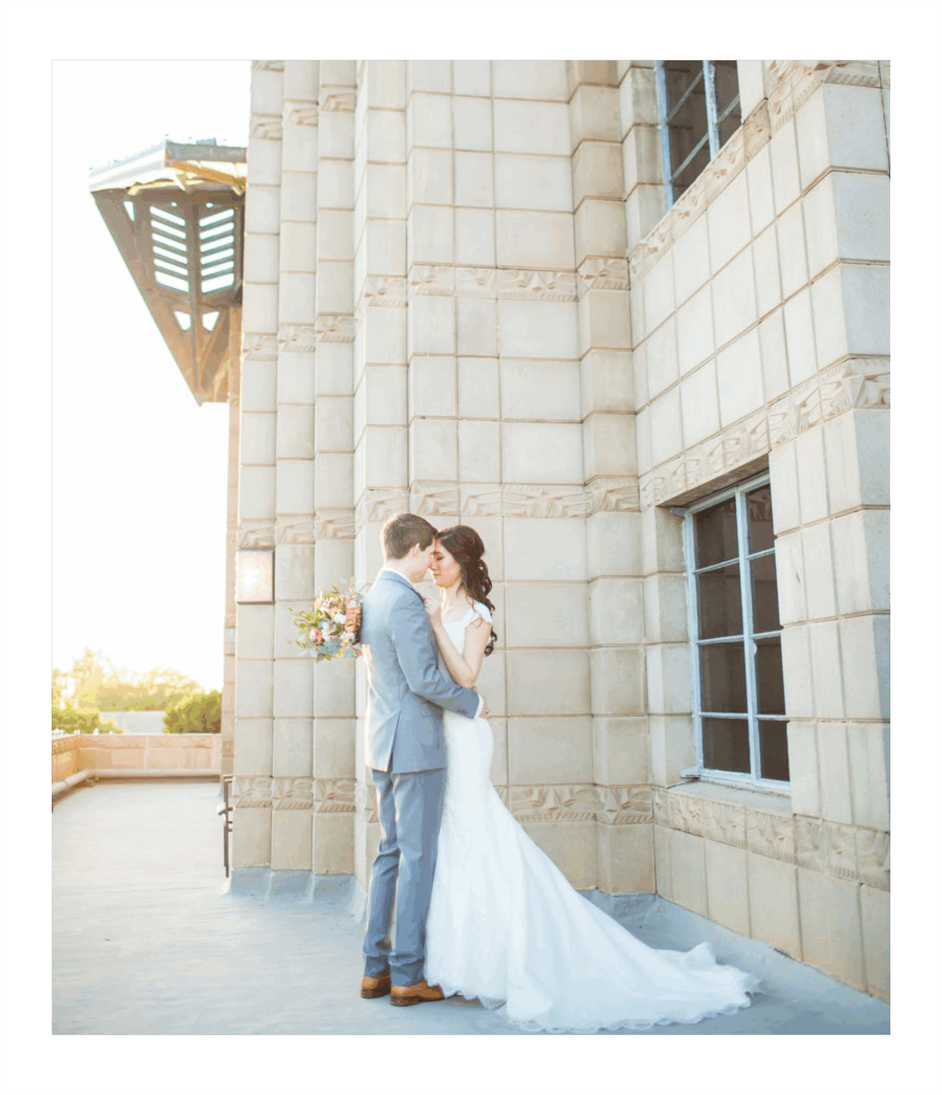 Bride and groom embrace by building wall.