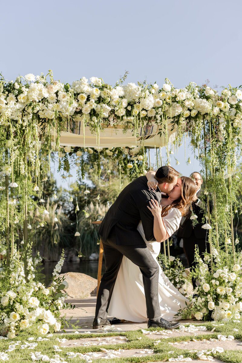 Bride and groom kissing under floral arch.