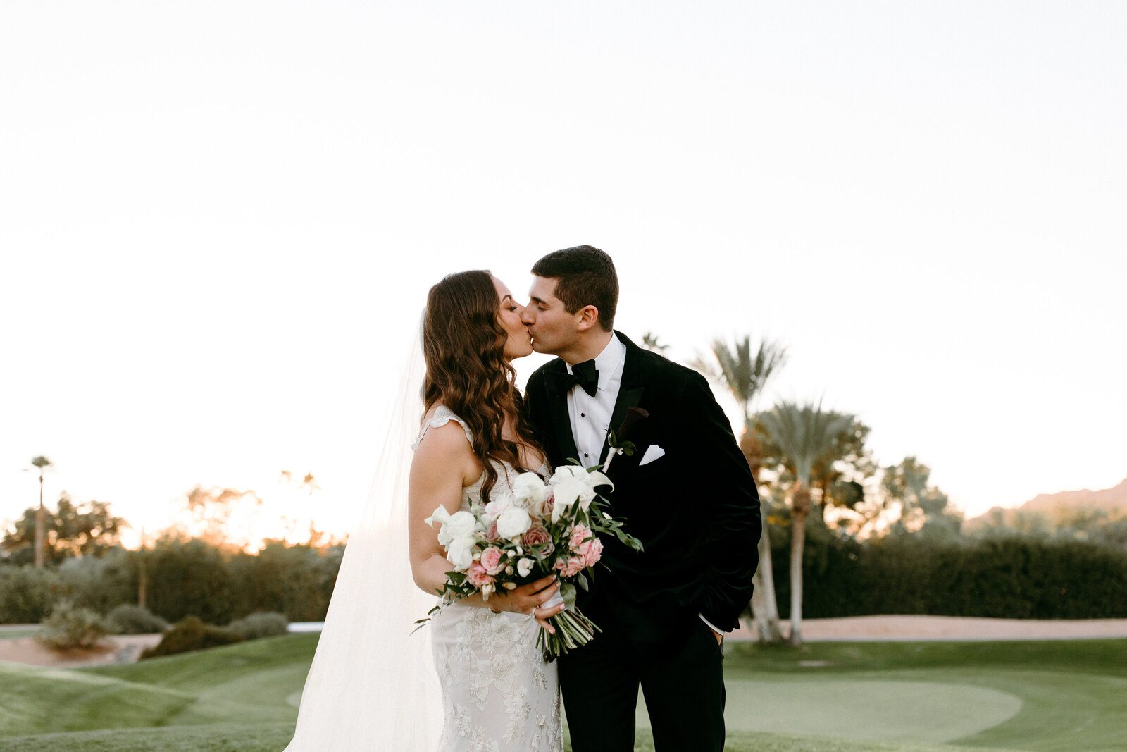 Bride and groom kissing outdoors, holding flowers.